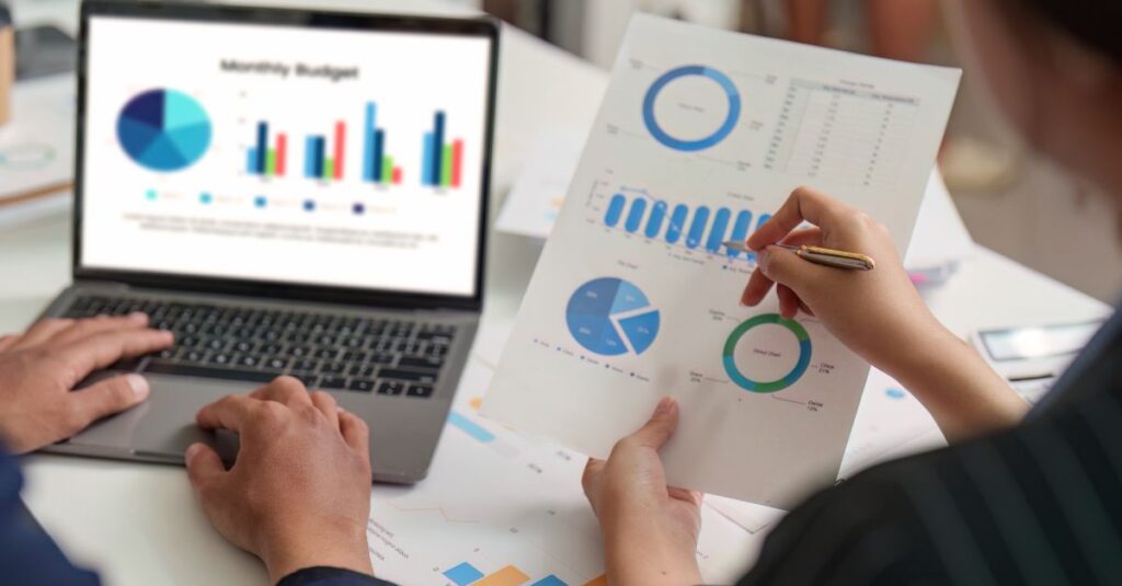 Two people analyze financial graphs and charts on a laptop and printed paper, reviewing Monthly Budget bar and pie charts in a business setting, utilizing CFO reporting tools for enhanced insights.